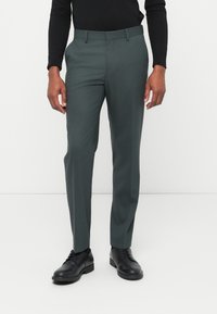 Σκούρο πράσινο παντελόνι με τελειώματα, με ομαλή υφή, διαθέτει επίπεδη μπροστινή όψη, θηλιές για ζώνη και στενή εφαρμογή, συνδυασμένο με μαύρα παπούτσια.