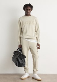 Sudadera beige con el texto "BOSS", joggers beige a juego con detalles de bolsillos, bolso negro y zapatillas blancas con suelas marrones.