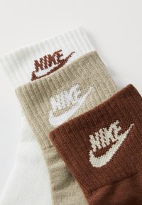 Tris porų Nike kojinių baltos, smėlio ir rudos spalvos. Kiekviena pora turi raukšlėtą kraštą ir Nike logotipą kontrastingomis spalvomis.