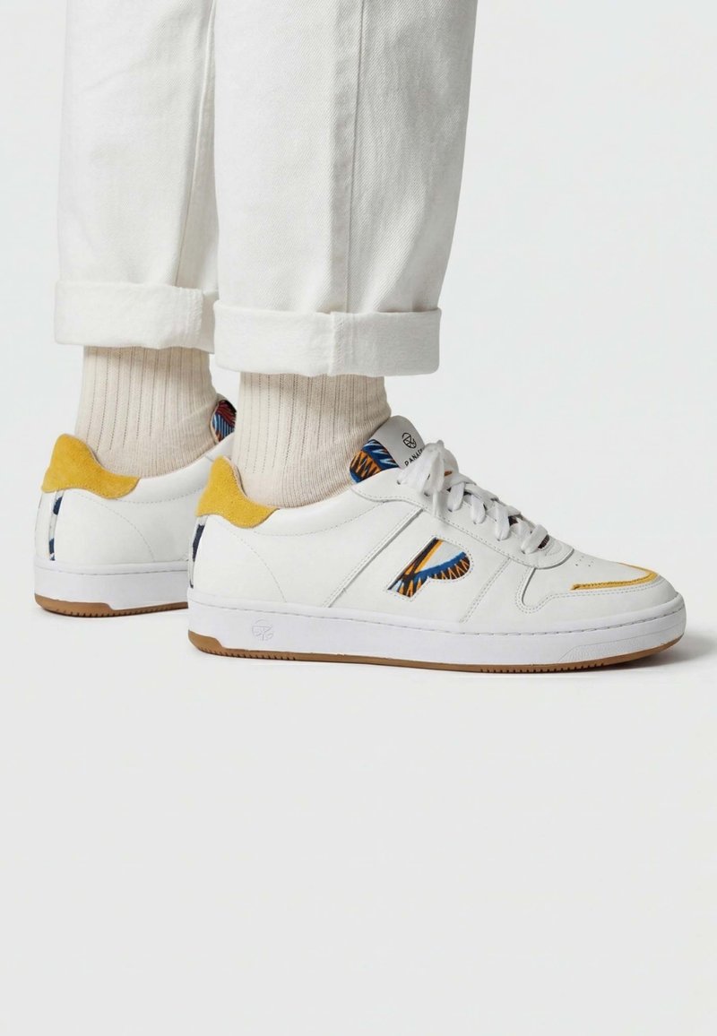 Baskets blanches avec des accents de talon en suède jaune et des détails bleus et oranges à motifs, portées avec des chaussettes crème et un pantalon blanc retroussé.