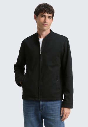 Man with dark hair wearing a black zip-up jacket over a white shirt and blue jeans, standing with one hand in pocket.