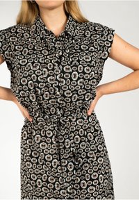 Vestito nero con bottoni, maniche corte e un motivo floreale circolare in beige e bianco. Include una vita legata per la regolazione.