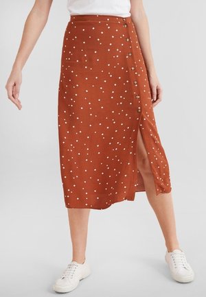 Jupe midi couleur rouille à pois blancs, détail boutons sur le côté et fente, portée avec des baskets blanches et un haut blanc.