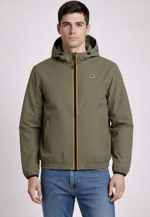 Młody mężczyzna w oliwkowo-zielonej kurtce z kapturem i przednim zamkiem błyskawicznym oraz niebieskich jeansach, stojący przodem na jednolitym tle.