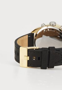 Correa de reloj de cuero negro con cortes cuadrados, hebilla de metal dorado y botones de crono en plata. Presenta una caja de reloj en tono dorado.