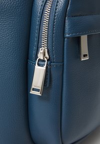 Mochila de piel azul con una superficie texturizada, herrajes de cremallera plateados y una etiqueta de marca. El diseño incluye bordes estructurados.
