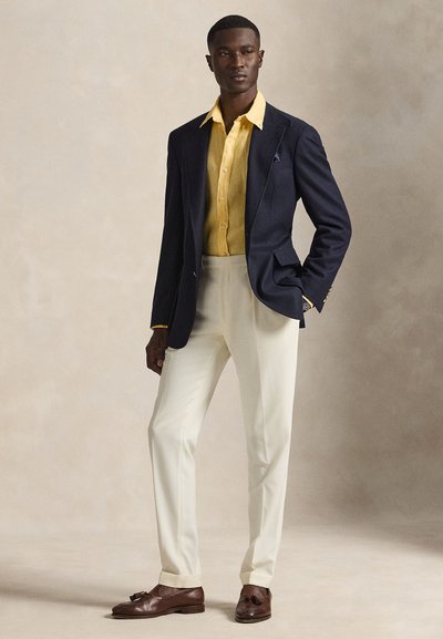 Σκούρο μπλε σακάκι ναυτικού τύπου, κίτρινο πουκάμισο, παντελόνι σε κρεμ απόχρωση και καφέ μοκασίνια. Ομαλές υφές, ραμμένη εφαρμογή και διακριτικές μεταλλικές λεπτομέρειες.