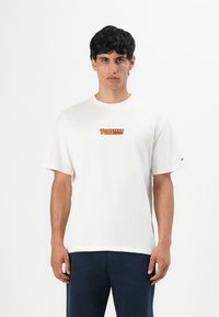 Man with dark hair wearing white Tommy Jeans t-shirt with orange logo and dark blue pants, standing against plain white background.