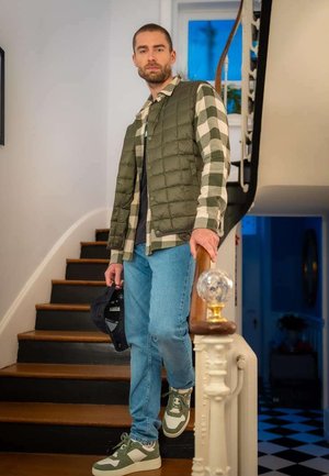Homme portant un gilet matelassé vert olive sur une chemise à carreaux, un jean bleu clair et des baskets vertes, se tenant sur un escalier.