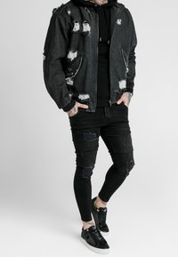 Czarna kurtka dżinsowa z postrzępionymi łatami, zestawiona z czarną bluzą z kapturem i podartymi czarnymi dopasowanymi dżinsami. Obuwie to czarne sneakersy z akcentami.