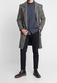 Abrigo gris texturizado, camisa azul marino con lunares, pantalones oscuros y botas negras con cordones y suelas marrones. Enfoque principal en el conjunto en capas y los contrastes.