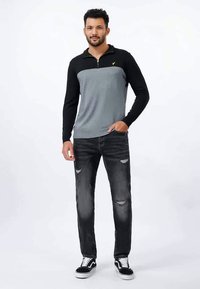 Melns un pelēks džemperis ar rāvējslēdzēja apkakli un ribotu tekstūru, valkāts kopā ar nodilušām tumšām džinsa biksēm un melnām sporta kurpēm ar baltiem zolēm.