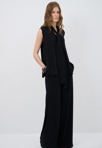 Top sans manche noir avec un décolleté noué, design superposé et coupe ample, associé à un pantalon noir à jambes larges, avec poches latérales.