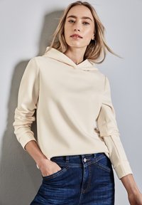 Hoodie beige chiaro realizzato in morbido tessuto, con tasca a marsupio sul davanti e polsini a coste, abbinato a jeans blu scuro.