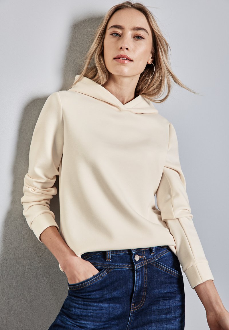 Hoodie beige chiaro realizzato in morbido tessuto, con tasca a marsupio sul davanti e polsini a coste, abbinato a jeans blu scuro.