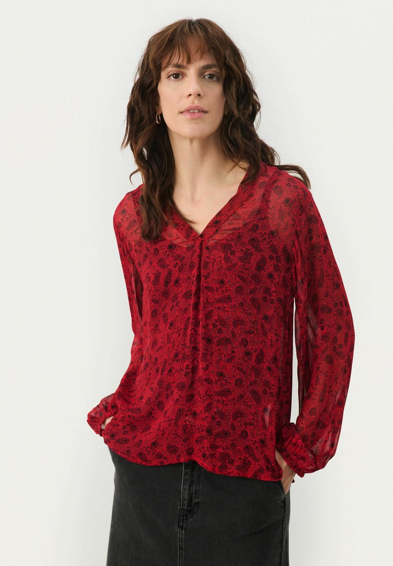 Blusa roja transparente con un patrón floral, escote en V y mangas largas. Presenta puños fruncidos y un corte holgado, combinada con una falda negra.