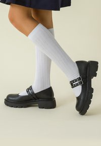 Zapatos Mary Jane de cuero negro con suela gruesa, que cuentan con dos hebillas ajustables y tachuelas decorativas. Combinados con calcetas blancas de canalé hasta la rodilla.