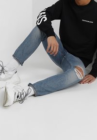 Person seated on floor wearing ripped blue jeans, white socks, chunky white sneakers, and a black sweatshirt with "Calvin Klein Jeans" text.