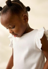 Top bianco a righe con maniche a sbuffo, indossato da un bambino con capelli ricci, che guarda in basso su uno sfondo morbido.