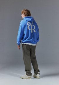 Jeune homme portant un sweat à capuche bleu avec un grand logo blanc "BR" dans le dos, pantalon gris, et baskets blanches, debout devant un fond gris.