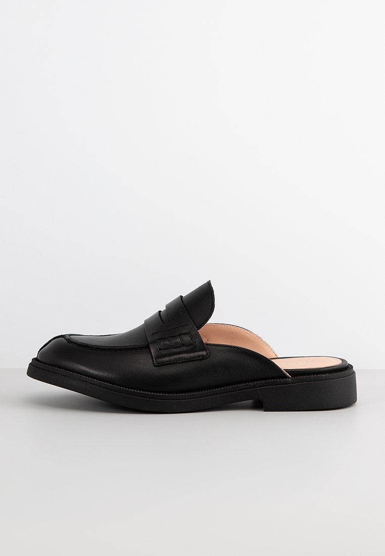 Schwarze Leder-Mules mit geschlossener Zehe, dekorativem Riemen und strukturiertem Detail. Mit rutschfester Gummisohle und glatter Innenausstattung.