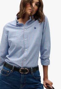 Lichtblauw overhemd met roze strepen, voorzien van een kraag en een geborduurd logo, gecombineerd met donkerblauwe denim en een zwarte riem.