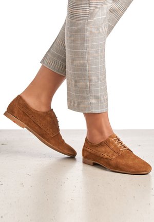 Braune Wildleder-Schnürschuhe mit perforierten Details und flacher Sohle, kombiniert mit grauen karierten Hosen, auf einem hellen strukturierten Hintergrund.