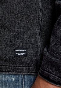 Veste en denim noir avec tissu texturé, coutures cousues, et une étiquette carrée portant les inscriptions "JACK & JONES" et "QUALITY ESSENTIALS".