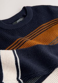 Pull en tricot avec une base bleu marine et des motifs texturés. Des rayures orange et grises ajoutent du détail, complétées par des accents crème et bleu marine.
