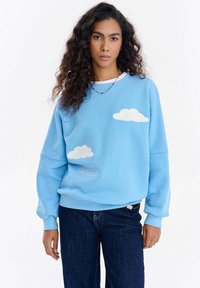 Jasnoniebieska bluza z miękkiego materiału, ozdobiona białym haftem w kształcie chmurki oraz nadrukowanym napisem u dołu. Luźny krój z ściągaczami na rękawach.