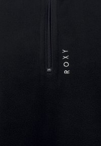 Tissu en polaire noire avec fermeture éclair noire centrale, texte blanc brodé "ROXY" positionné verticalement à droite de la fermeture éclair.
