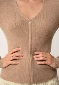 Cárdigan de punto acanalado color beige con escote en V y cremallera de media longitud. Textura suave, diseño ajustado y mangas largas acanaladas, usado sobre una falda plisada.