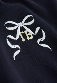 Tela azul marino con un lazo blanco bordado y las iniciales doradas "TB" en el centro, mostrando una textura suave y un diseño limpio.