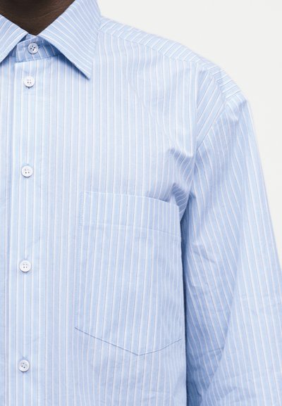 Ανοιχτό μπλε πουκάμισο με λευκές κάθετες ρίγες, λευκά κουμπιά, τσέπη στο στήθος και μυτερό γιακά.