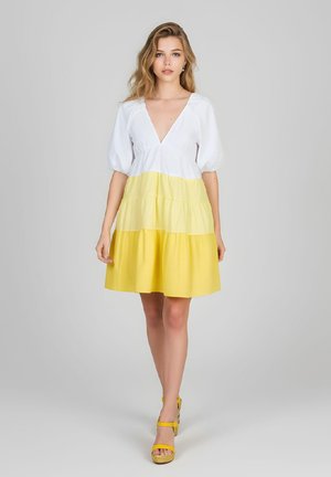Mujer con un vestido de volantes blanco y amarillo bicolor con mangas abullonadas caminando hacia adelante, combinado con sandalias amarillas de plataforma.
