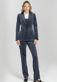Kobieta stoi, ubrana w granatowy, kordurowy garnitur z marynarką, dopasowanymi spodniami, białą koszulą i czarnymi butami na obcasach na szarym tle.