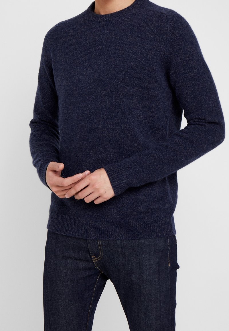 Maglione di lana blu navy con collo rotondo, polsini e fondo a costine. Design aderente, lavorazione a maglia strutturata, indossato sopra jeans scuri.