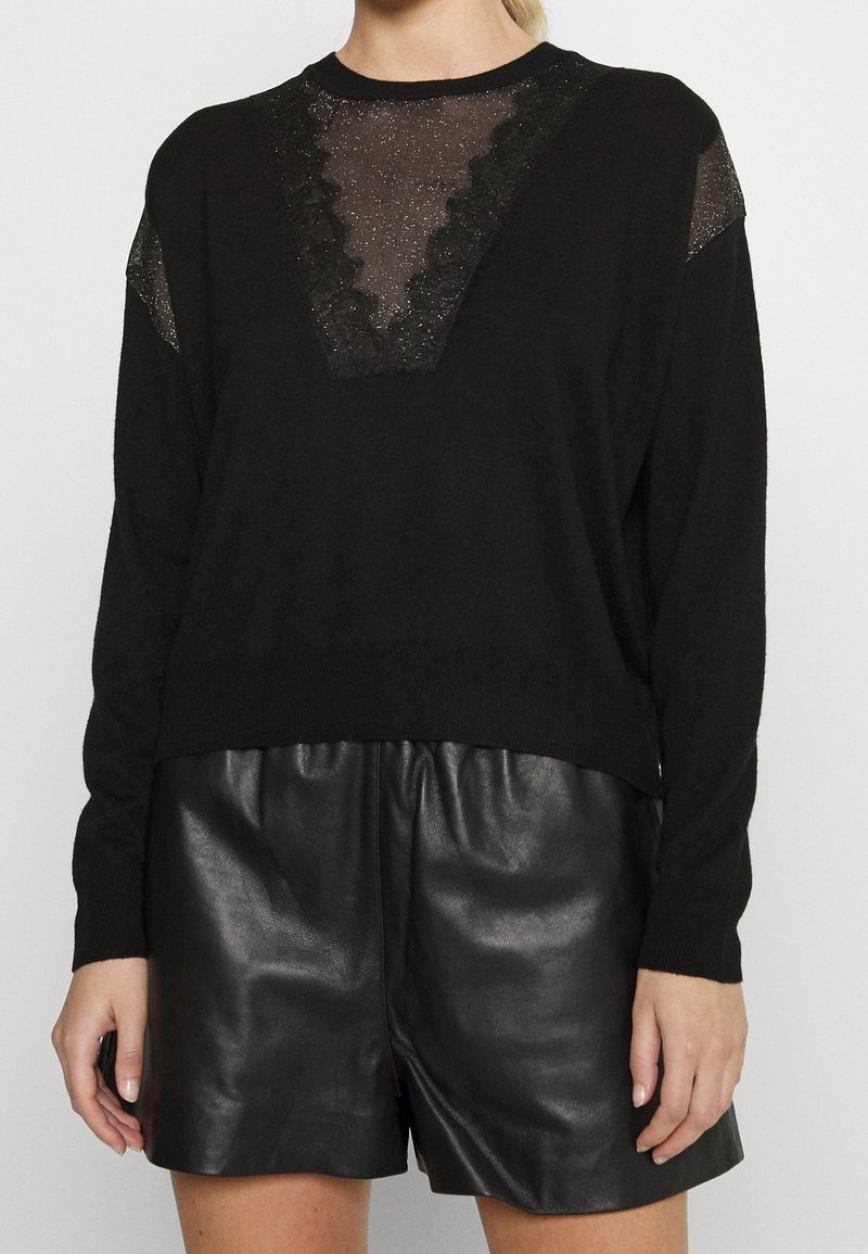 Pull noir à manches longues avec un col en V en dentelle transparente scintillante et des panneaux aux épaules, associé à un short en cuir noir sur une personne debout.