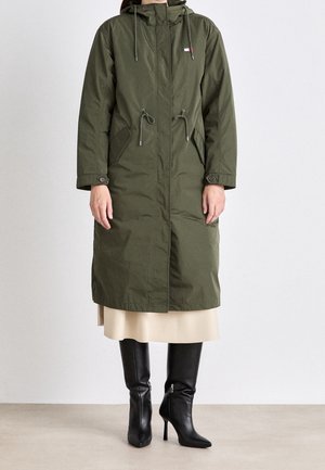 Femme portant un long parka vert olive avec cordons et poches avant, jupe beige et bottes noires à talons montant jusqu'au genou.