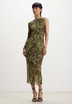 Vestito verde oliva a spalla unica con silhouette aderente, caratterizzato da motivi astratti e una parte inferiore trasparente, abbinato a tacchi neri con cinturini.