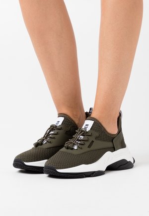 Pieds portant des baskets en maille vert olive avec semelles blanches et semelles extérieures noires, dotées de lacets élastiques à cordon et de languettes de traction blanches.