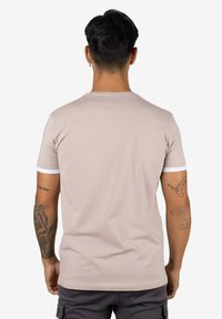 Camiseta beige de manga corta con detalles blancos en los puños y el cuello. Tela suave. Se ve la espalda de una persona con tatuajes.
