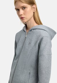 Szara bluza zapinana na zamek z miękką fakturą, wyposażona w dużą kapturem, boczne kieszenie oraz subtelnie haftowane logo na piersi.