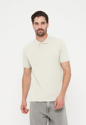 Uomo con capelli scuri e barba che indossa una polo beige chiaro e jeans grigi in piedi davanti a uno sfondo bianco semplice.