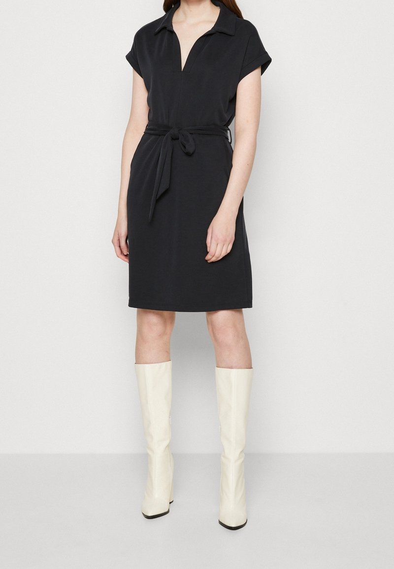 Robe noire à manches courtes avec col et taille ceinturée. Tissu lisse, longueur genou, associée à des bottes blanches brillantes montant jusqu'au genou.