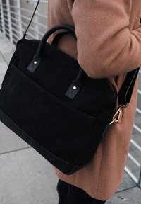 Black canvas tote bag with leather handles, featuring a front pocket, gold hardware accents, and a structured design.