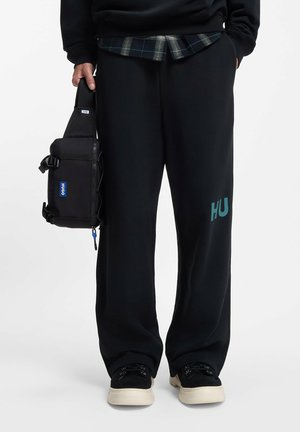 Person wearing black wide-leg pants with "HU" logo, black sneakers with white soles, holding a black rectangular bag with "Hugo" label.