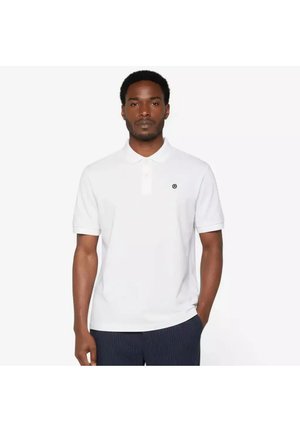 Man staat met het gezicht naar voren gericht, draagt een wit poloshirt met korte mouwen en een klein cirkelvormig logo op de borst, en donkere gestreepte broek.