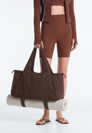 Personne en tenue de sport marron tenant un grand sac cabas marron avec un tapis de yoga blanc roulé attaché en dessous.
