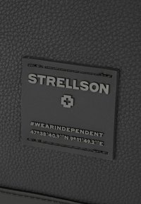 Črn usnjen obliž z teksturo, ki prikazuje logotip "STRELLSON", haštag "#WEARINDEPENDENT" ter koordinate gravirane na spodnjem delu.